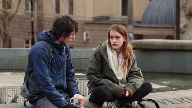 Zwei junge Menschen sitzen auf einer steinernen Umrandung, einer hält eine Tasse, beide wirken nachdenklich.