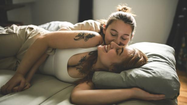 Zwei Frauen liegen lächelnd im Bett, eine gibt der anderen einen Kuss auf die Wange.