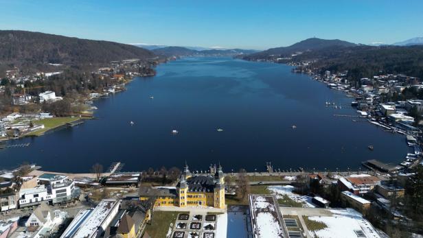 Ein gelbes Schloss liegt an einem See, umgeben von Bergen und Gebäuden.