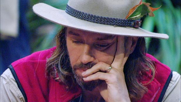 Ein Mann mit langem Haar und Hut - Gil Ofarim - stützt nachdenklich sein Gesicht auf die Hand.