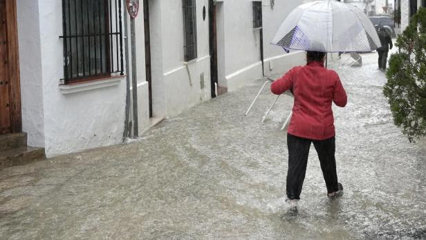 Eine Person mit Schirm watet durch eine überflutete Gasse mit weißen Häusern.