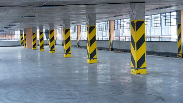 An expansive parking garage features yellow and black safety barriers on the columns, illuminated by natural light from large windows