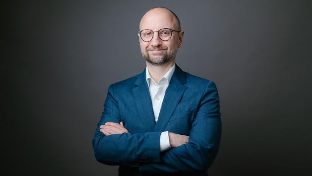 Mann mit Brille im blauen Anzug steht mit verschränkten Armen vor grauem Hintergrund.