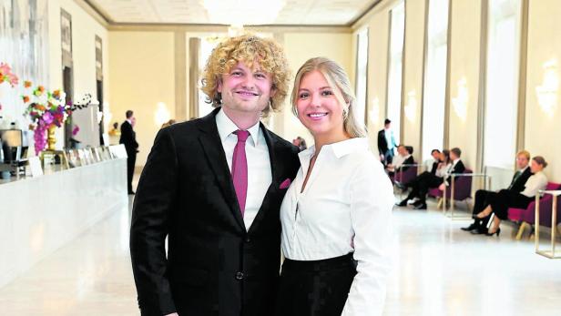 Ein Mann im Anzug und eine Frau im eleganten Outfit stehen lächelnd in einem hellen, prunkvollen Saal.