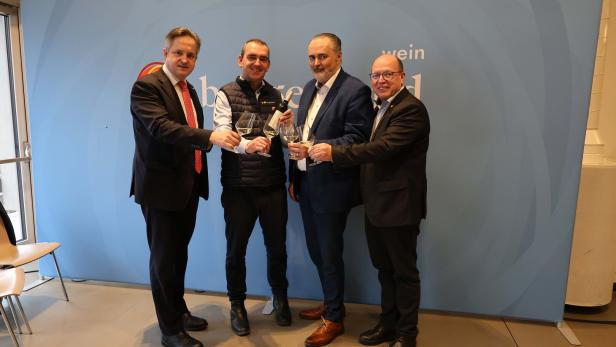 Vier Männer stoßen mit Weingläsern an und stehen vor einer blauen Wand mit Flaschen auf einem Regal darüber.