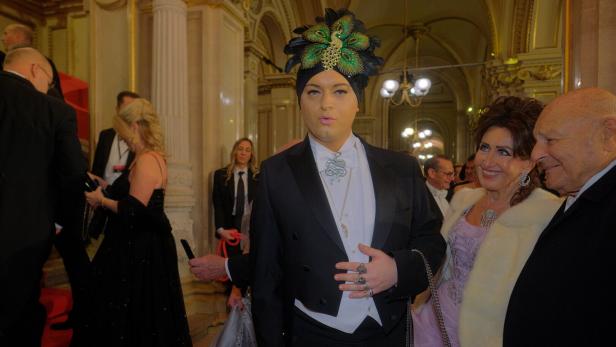 Ein Mann mit Turban und Pfauenfedern posiert mit elegant gekleideten Personen.