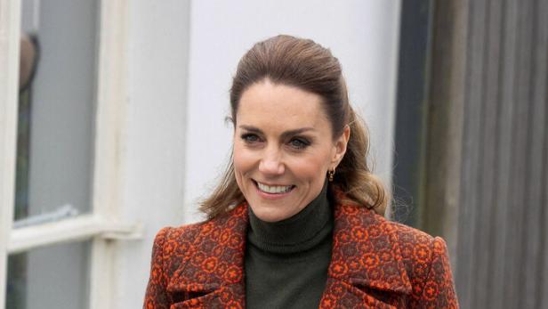 Prinzessin Kate mit braunen Haaren trägt einen gemusterten orange-roten Mantel und einen dunkelgrünen Rollkragenpullover.