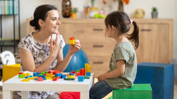 Eine Frau zeigt einem Mädchen mit Bauklötzen spielerisch etwas und deutet dabei auf ihren Mund.