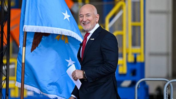 Ein Mann im Anzug und mit roter Krawatte vor einer blauen Flagge