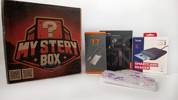 Mystery Box mit Elektronikzubehör, Kopfhörern, Kartenleser und Handyhülle auf weißem Hintergrund.