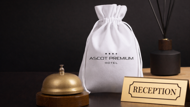 Weißer Stoffbeutel mit Hotellogo neben goldener Klingel und Rezeption-Schild auf dunklem Tisch.