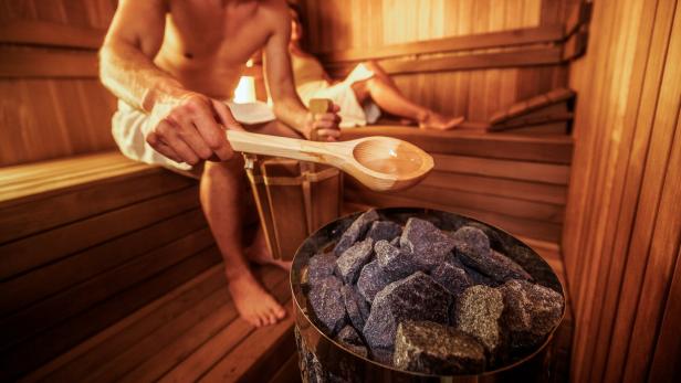 Zwei Menschen sitzen umschlungen von Handtüchern in einer Sauna, der Mann im Zentrum des Bilds schöpft Wasser auf die heißen Steine.