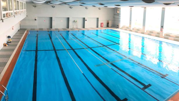 Leeres Hallenbad mit mehreren Schwimmbahnen und Tageslicht durch große Fenster.