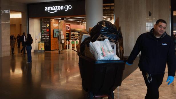 Ein Mann schiebt einen großen Müllwagen durch eine Einkaufspassage vor einem Amazon-Go-Laden.