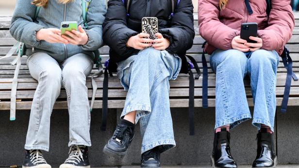 drei Mädchen sitzen auf einer Bank nebeneinander und schauen aufs Handy, ihre Köpfe sind abgeschnitten