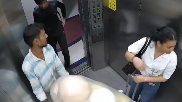 Drei Personen stehen in einem Aufzug, eine Frau mit Koffer und zwei Männer, einer davon hält mehrere Luftballons in der Hand.