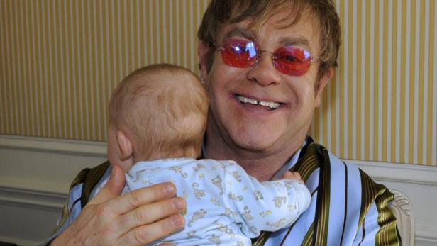 Elton John hält ein Baby im Arm und lächelt.