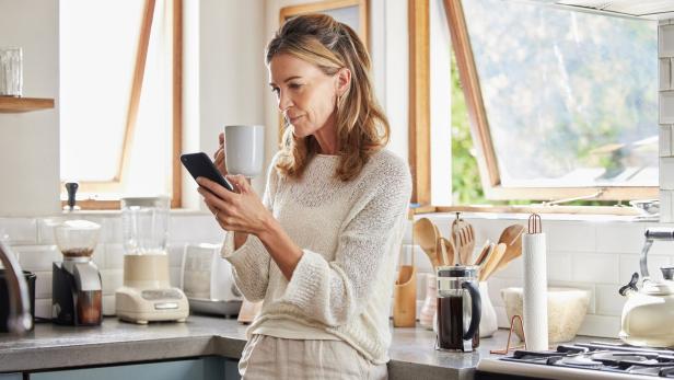 Coffee, phone and senior woman in kitchen browsing social media, text message or online app at home. Relax, tea and reading news or mobile online web surfing with female from Canada on 5g smartphone