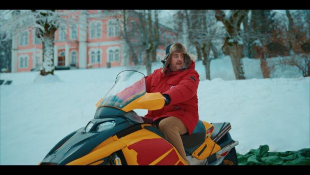 Bastian Pastewka in roter Winterjacke und Fellmütze sitzt auf einem gelb-schwarzen Schneemobil vor einem verschneiten Gebäude.