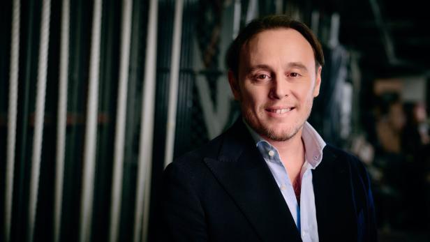 Ein lächelnder Michele Mariotti mit Sakko und hellblauem Hemd vor einem Hintergrund mit vertikalen Streben.