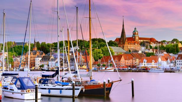 Segelboote liegen im Hafen von Flensburg vor der Kulisse der Altstadt mit historischen Gebäuden und der St.-Nikolai-Kirche bei Sonnenuntergang.