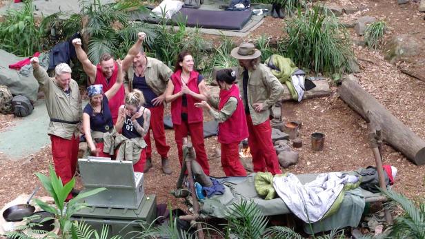 Mehrere Dschungelcamp-Teilnehmer jubeln gemeinsam im Camp zwischen Pflanzen und Schlafplätzen.