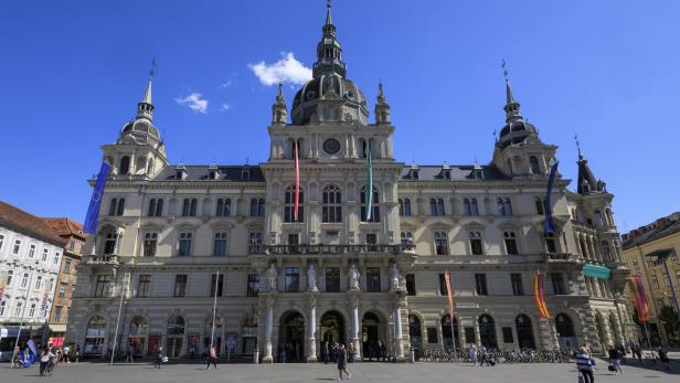 Das Grazer Rathaus präsentiert sich mit Fahnen und belebtem Vorplatz unter blauem Himmel.