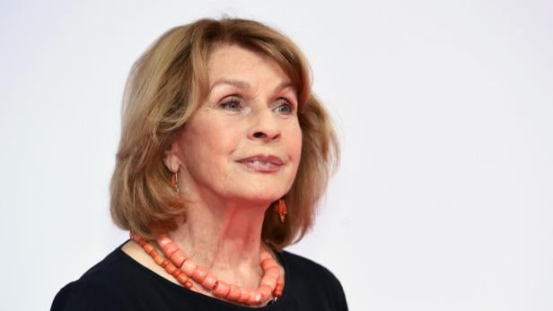 Eine Frau mit blondem Haar trägt eine korallenfarbene Halskette und Ohrringe vor einem hellen Hintergrund.