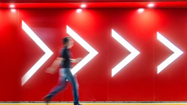 Eine Person geht vor einer roten Wand mit vier großen, leuchtenden Pfeilen nach rechts entlang.