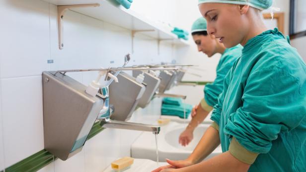 Zwei Personen in OP-Kleidung waschen sich gründlich die Hände an einem Waschbecken mit Seifenspendern.
