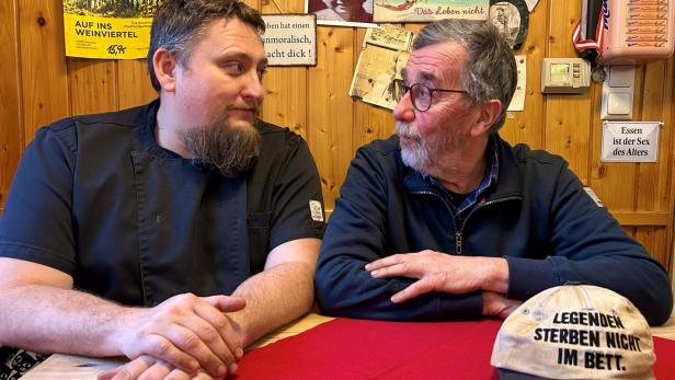 Zwei Männer sitzen an einem Tisch mit roter Decke, im Hintergrund Holzvertäfelung mit Dekoration.