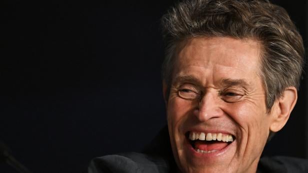 Der Schauspieler Willem Dafoe, hellauf lachend vor dunklem Hintergrund 