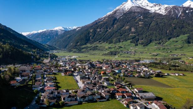 Ein Dorf liegt in einem Tal, umgeben von grünen Hängen und schneebedeckten Bergen.