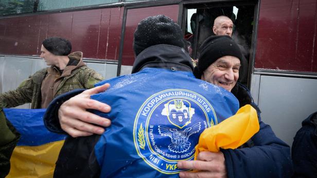Ein Mann umarmt eine Person mit einer blauen Jacke mit ukrainischem Wappen vor einem Bus.