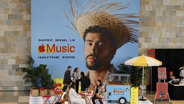 Mehrere Menschen sitzen an roten Tischen vor einer großen Werbewand für den Super Bowl LX Apple Music Halftime Show.