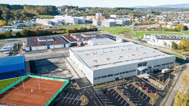 Luftaufnahme eines Gewerbegebiets mit Hallen, Tennisplätzen, Parkplätzen, Sportfeldern und Wohnhäusern im Hintergrund.