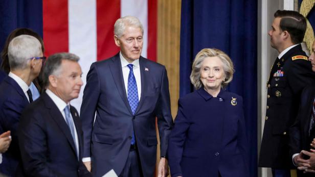 Bill und Hillary Clinton stehen vor einer US-Flagge und anderen Personen.
