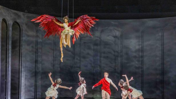 Eine Person mit großen roten Flügeln schwebt über vier tanzenden Menschen und einer stehenden Person auf einer Theaterbühne.