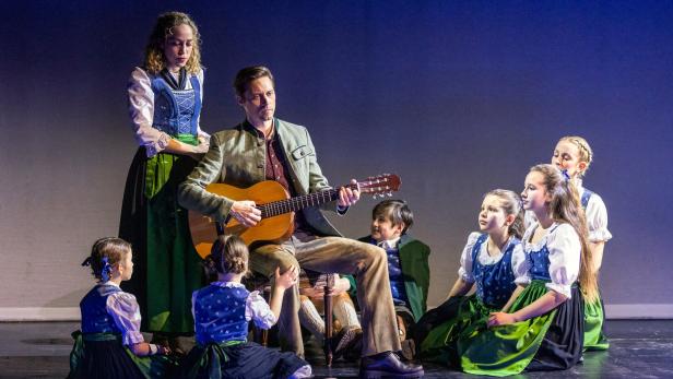 Ein Mann spielt Gitarre, umgeben von Kindern und einer Frau in traditionellen Trachten auf einer Bühne.