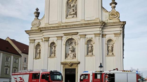 Feuerwehrfahrzeuge stehen vor der Kirche in Bruck an der Leitha, während Einsatzkräfte nach dem Brand am Eingangsbereich tätig sind.