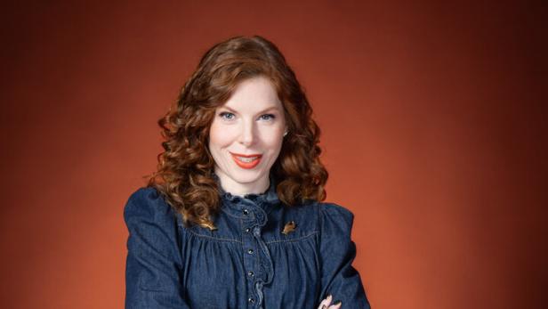 Frau mit lockigen, roten Haaren trägt eine dunkelblaue Jeansbluse und Jeanshose vor braunem Hintergrund.