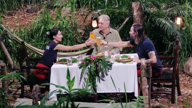 Am festlich gedeckten Tisch mitten im Dschungel stoßen Samira Yavuz, Hubert Fella und Gil Ofarim auf die Dschungelzeit an.