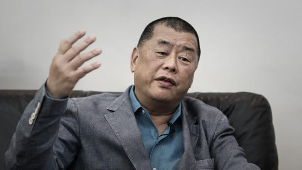 Ein Mann im grauen Sakko sitzt auf einem dunklen Sofa und gestikuliert mit der rechten Hand.