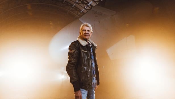 Ein älterer Mann in Fliegerjacke steht in einer beleuchteten Halle vor einem großen Luftschiff.