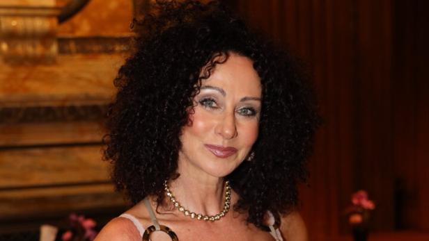 Frau mit lockigen dunklen Haaren trägt ein goldenes, glänzendes Kleid und Schmuck, steht in elegantem Innenraum.