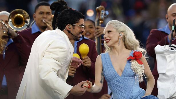 Ein Mann im weißen Anzug und eine Frau im blauen Kleid halten Händchen und lächeln sich auf einer Bühne mit Musikern an.