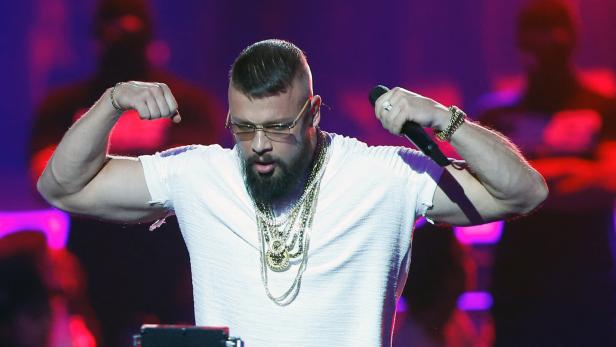 Ein Mann mit Bart und Goldketten zeigt seine Muskeln auf einer Bühne mit buntem Licht.