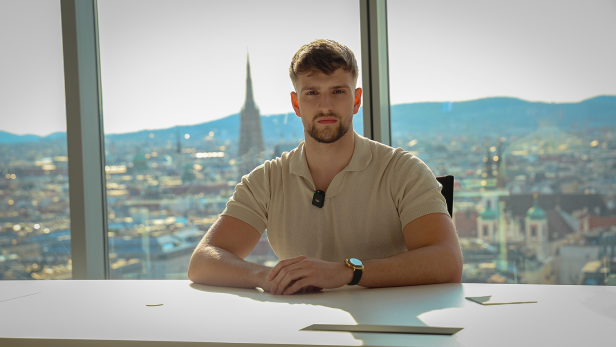 Kilian Kropiunik sitzt an einem Schreibtisch vor der Wiener Skyline und blickt ernst in die Kamera.