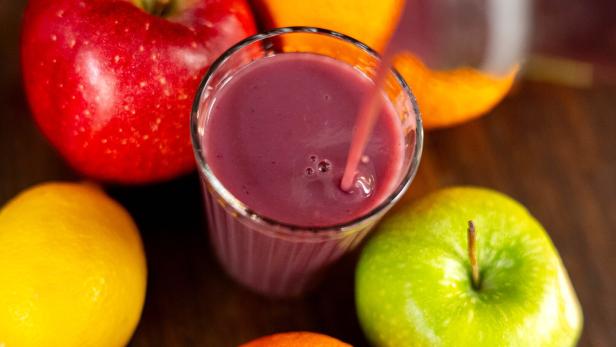 Ein Glas lila Smoothie wird umgeben von Apfel, Orange und Zitrone eingeschenkt.