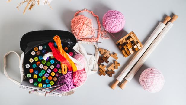 Analog bag of yarn, markers, a notebook, knitting needles, and puzzles top view web banner